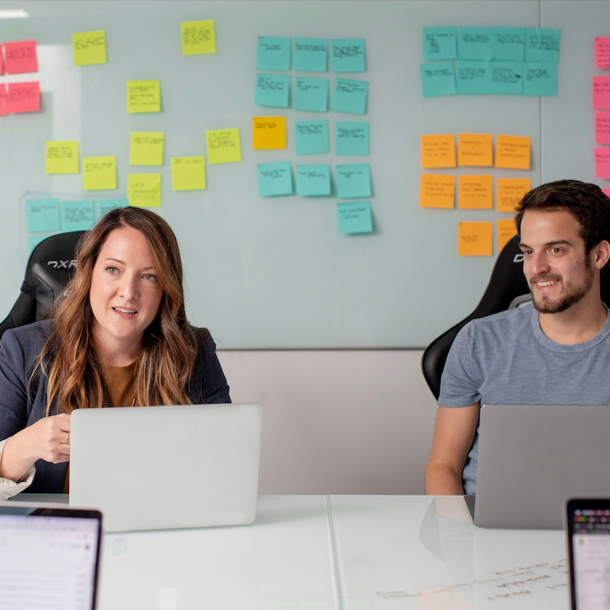 Two employees sit at a table with laptops. Sticky notes are on the wall behind them.